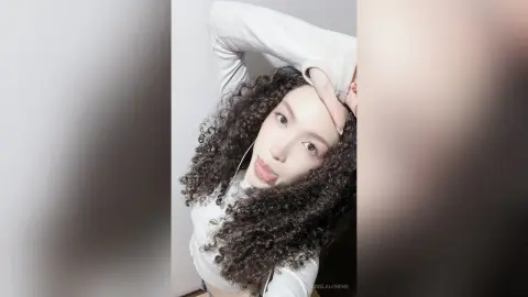 Young woman with curly dark hair wearing a white long-sleeve top in a soft, minimalist indoor setting.