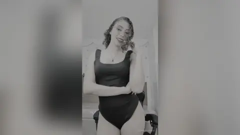 Smiling woman in a black bodysuit posing in a bright bedroom setting.