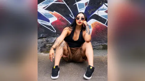 ChloeRiveiro — Tattooed woman in black tank top and brown shorts sitting against colorful graffiti wall in a cool urban mood.