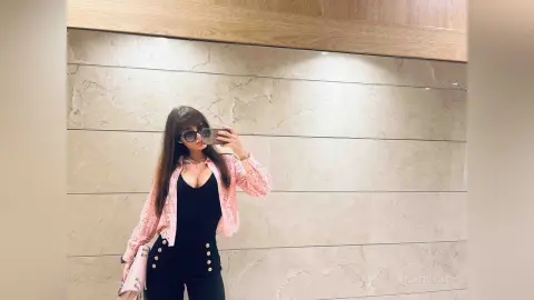 VesperMuse poses in a mirror wearing a pink button-up shirt over a low-cut black tank top and black pants, showing cleavage and her long brown hair.