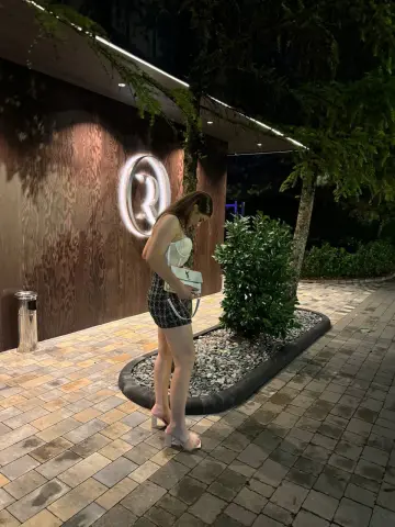 Woman in a white top and plaid shorts standing by a modern wood-paneled building at night.