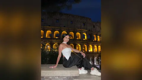 Stylish woman in a white halter top and black trousers sitting before the illuminated Roman Colosseum at night.