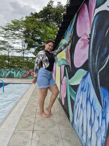 Smiling woman in a long-sleeve swim top and denim shorts posing by a colorful floral mural next to a swimming pool.