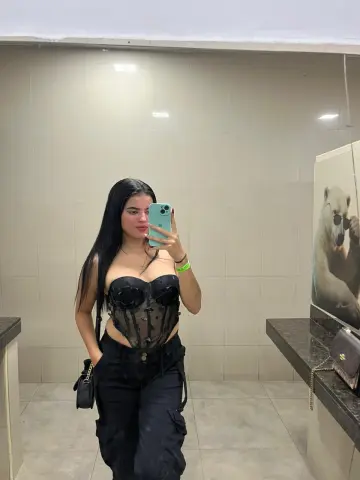 Young woman in black corset top and cargo pants taking a mirror selfie in a modern bathroom.