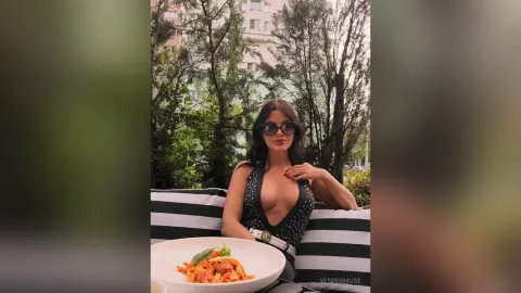 VesperMuse wearing black sunglasses and a low-cut black top, sitting outdoors behind a plate of pasta.