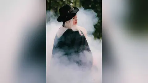 EllieFrosty wears a black hat and a dark cloak while standing amidst thick white smoke or fog.