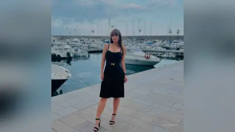 VesperMuse stands on a stone pier wearing a black sleeveless dress and black ankle-strap heels, with a harbor full of white boats behind her.