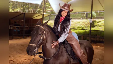 Stylish woman in western cowgirl attire riding a dark brown horse in a rustic stable setting.