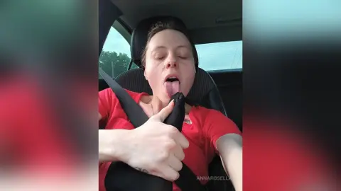Woman in a red shirt sticking her tongue out playfully while sitting in a car seat with a seatbelt.