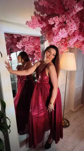 Elegant woman in a shimmering burgundy evening gown posing by pink cherry blossom decor in a bright room.