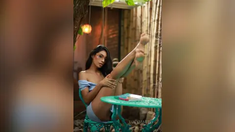 LucyMartin sits on a teal chair outdoors, wearing a light blue off-the-shoulder top while pulling her bare legs up toward her chest.