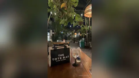 Black Marc Jacobs tote bag sitting on a wooden cafe table in a lush, dimly lit outdoor garden setting.