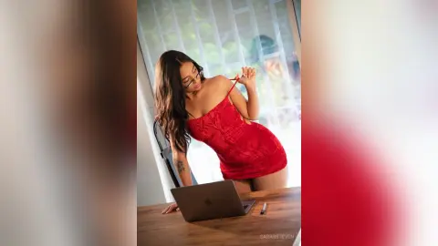 Glamorous woman in a red cocktail dress posing by a laptop in a bright, modern indoor setting.