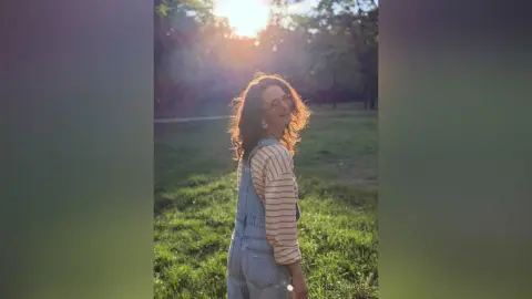 Smiling woman in striped shirt and denim overalls standing in a sunlit green meadow at golden hour.