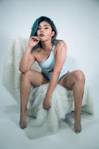 Tattooed woman in a light blue bodysuit sitting on a white faux fur rug against a clean studio background.