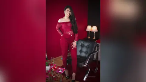 SamanthaHarvey stands in a red lace off-the-shoulder top and matching red pants, posing next to a black leather chair.
