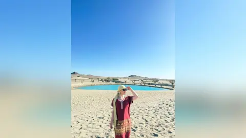 Woman in a maroon patterned tunic and headscarf standing in a sunny desert oasis by a blue lagoon.
