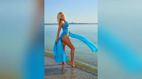 Anastasia stands on a sandy beach wearing a blue cut-out swimsuit with a long flowing skirt, posing against the ocean waves.