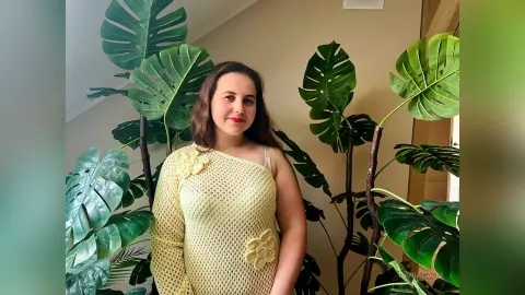 DariaCalm poses in a yellow knit one-shoulder top, standing in front of large green monstera leaves.