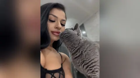 AlmaKane — Beautiful woman in a black lace top sharing a sweet, affectionate moment with her grey cat indoors.
