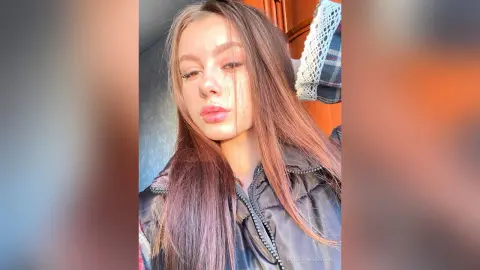 Young woman with long brown hair wearing a black puffer jacket in warm, natural sunlight.