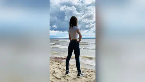 Young woman in white t-shirt and blue jeans standing on a sandy beach under a cloudy sky.