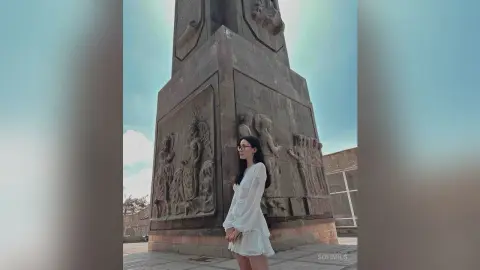 SofiMils stands outdoors next to a large stone monument, wearing a white long-sleeved blouse and glasses with her long dark hair down.