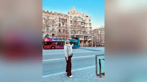 MilenaBia stands on a city street wearing a white jacket and dark maroon trousers next to a red Barcelona bus.