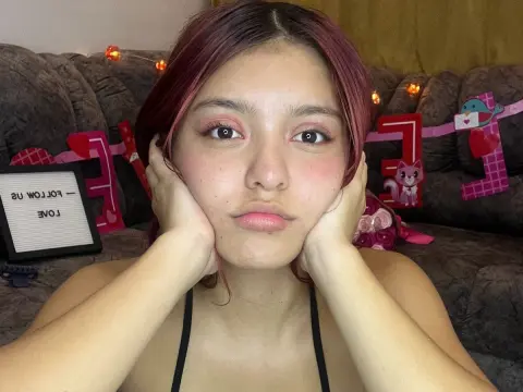 AlyMartini with reddish hair poses with her hands on her cheeks, wearing a black top while sitting on a couch.