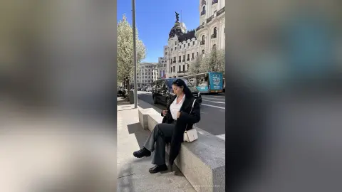 NashiroSakurai sits on a stone ledge in a black coat and grey trousers, looking down while holding a phone.