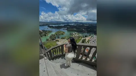 IvyStuart stands on a stone staircase overlooking a wide lake and green hills, wearing a black patterned top and white pants.