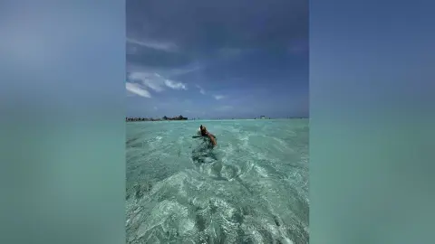 A person relaxing on a surfboard in turquoise tropical ocean water under a cloudy sky.