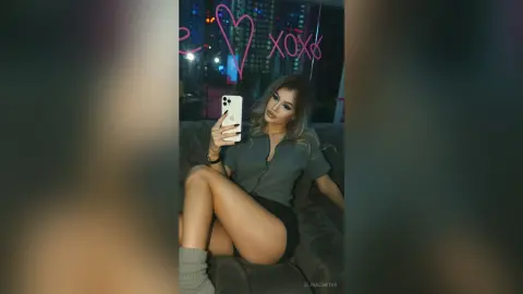 Stylish woman in a grey shirt taking a moody selfie on a couch in front of a pink neon heart sign.