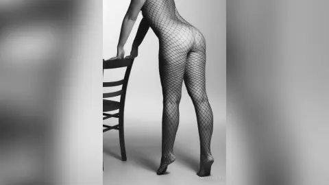 TannySmith poses in a black fishnet bodysuit, leaning forward over a chair against a plain background.