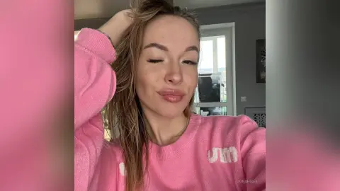 Smiling young woman in a cozy pink sweatshirt relaxing in a bright, sunlit room.