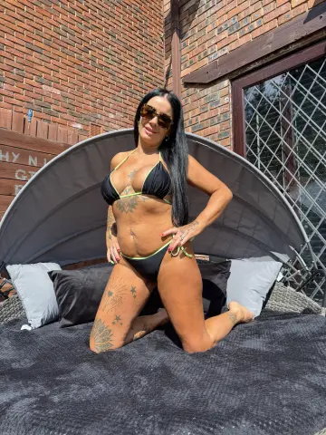 Tanned woman in black bikini kneeling on a lounge chair outdoors by a brick building in sunny weather.