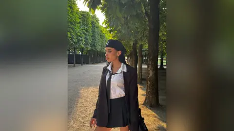 Stylish young woman in a black blazer and white shirt posing in a sunlit park with lush green trees.