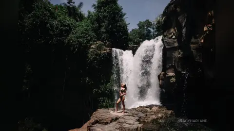 StacyHarland stands naked on a rock in front of a large, rushing waterfall surrounded by dense green jungle trees.