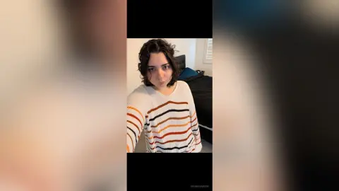 Young woman with wavy dark hair wearing a striped white and orange sweater in a cozy bedroom setting.