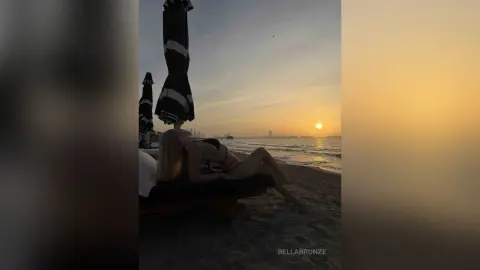 BellaBronze lies naked on a beach lounge chair at sunset, showing her long blonde hair and bare body against the ocean waves.