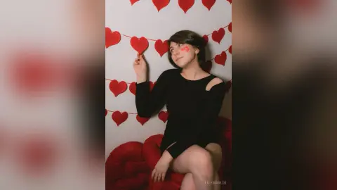 Young woman in a black long-sleeve top sitting by red heart decorations in a romantic indoor setting.