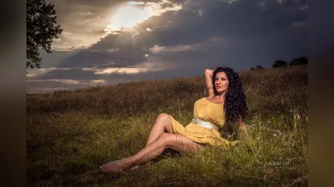 MissyJolie sits in a grassy field wearing a short yellow sundress with her long dark hair flowing down her back.