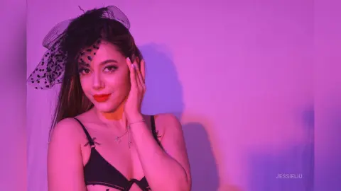 JessieLiu poses in a black lace bra with her hand touching her face against a purple background.