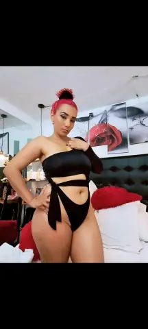 Curvy woman in a black cutout bodysuit posing confidently in a modern bedroom setting.