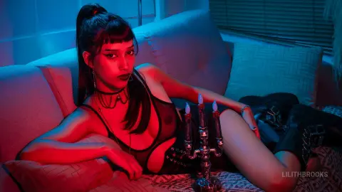 LilithBrooks reclines on a light sofa wearing a black sheer bodysuit and a black choker under dim red and blue lighting.