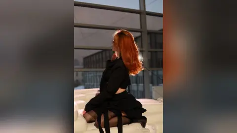 AlbertaCorrell is a redhead in a black outfit posing by a large window overlooking the city at night.