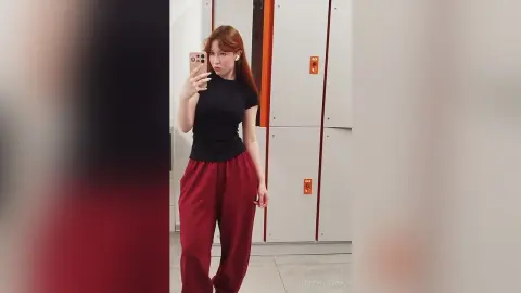 Redheaded woman in a black t-shirt and burgundy lounge pants taking a mirror selfie in a modern hallway.