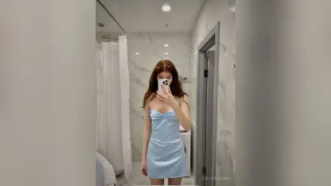 Young woman in a light blue satin slip dress taking a selfie in a modern marble bathroom.