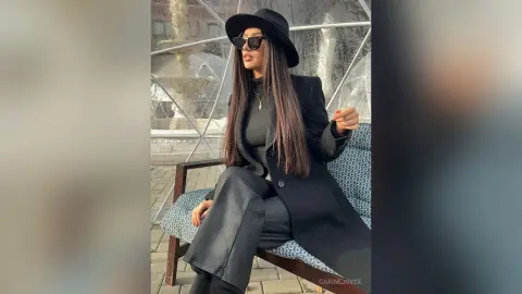 Carine wears a black hat, sunglasses, and a long black coat while sitting on a patterned chair outdoors.