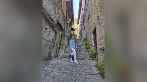 Stylish woman in a light blue jacket and white trousers walking down a rustic cobblestone alleyway in Europe.